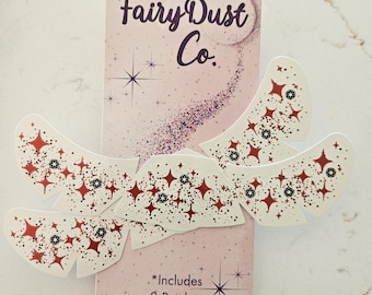 May include: A package of Fairy Dust Co. patches with a pink background and the text "Fairy Dust Co." in dark purple. The patches are white with red star and black dot designs. The package includes 2 patches.