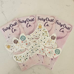 以下が含まれることがあります： 「Fairy Dust Co.」の文字と星柄が入ったピンク色のカードが複数枚。バラ、太陽、渦巻きなどのデザインが施された、星柄の白い切り抜きが上に配置されています。全体的なテーマは、気まぐれで魔法的です。