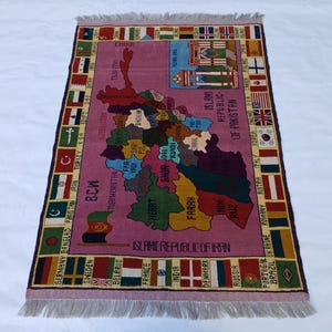 May include: A rectangular rug with a map design, featuring a central pink field with a detailed map of a region, surrounded by country flags. The rug has a fringed edge and text in multiple languages.