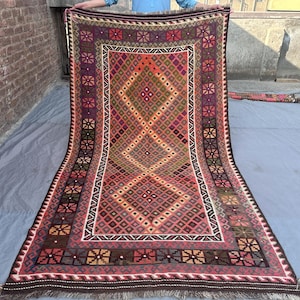Könnte beinhalten: Ein rechteckiger, handgewebter Teppich mit einem geometrischen Muster in Rot-, Lila-, Orange- und Brauntönen. Der Teppich hat ein zentrales Rautenmuster, das von einem Rand aus sich wiederholenden Blumenmotiven umgeben ist. Der Teppich hat einen Fransenrand.