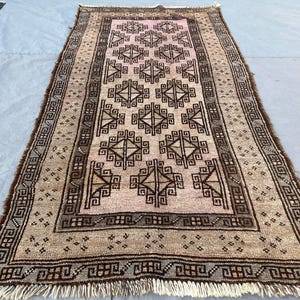 Puede incluir: Una alfombra rectangular, tejida a mano, con un campo central rosa y intrincados patrones geométricos en marrón. La alfombra tiene un borde con diseños geométricos repetidos y flecos en los extremos. La alfombra es probablemente de lana.