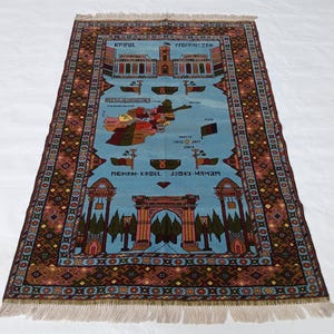 May include: A rectangular rug with a light blue central field featuring a map of Afghanistan and architectural illustrations. The rug has a brown border with geometric patterns and fringe on the short ends. The rug has text in the image.