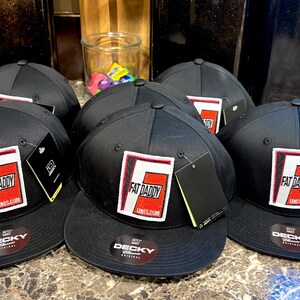 May include: A collection of black baseball caps with a flat brim. Each cap features a rectangular patch with the words "FAT DADDY" in a white and red design. The brand "DECKY" is visible on the brim.