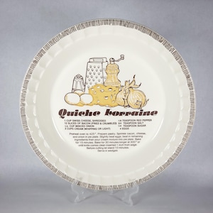 May include: A round, off-white ceramic pie dish with a fluted edge and brown striped rim. The center features a brown graphic of a grater, pepper mill, cheese, and onion, with the text Quiche Lorraine and a printed recipe for the dish.