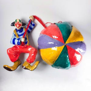 May include: A whimsical clown figurine with a red nose, painted face, and colorful outfit. The clown is pulling a large, multi-colored ball with a red rope. The clown wears red pants, yellow shoes, and a blue and white striped shirt.