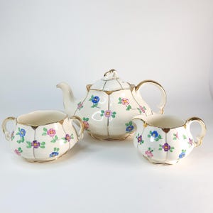 May include: A vintage tea set featuring a teapot, sugar bowl, and creamer. The set is cream-colored with gold trim and decorated with blue, pink, and purple floral patterns. The teapot has a lid and a curved handle.