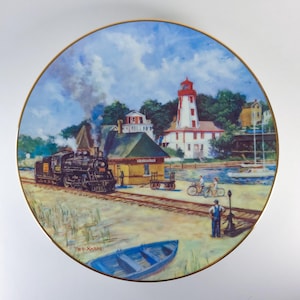 Peut inclure: Assiette décorative avec une illustration d'une gare, d'un phare et d'un train à vapeur. L'assiette a un bord doré et un bateau bleu au premier plan. Le texte "Kincardine" est visible sur la gare.