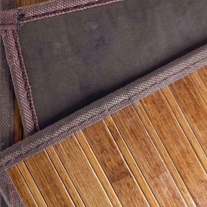 Puede incluir: Primer plano de una estera de bambú con un respaldo de tela marrón y un borde tejido. Las láminas de bambú son de color marrón claro y el borde es de un marrón más oscuro. La estera está diseñada para su uso como revestimiento de suelo.