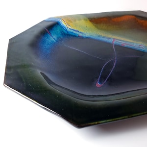 Puede incluir: Un plato de cerámica octogonal hecho a mano con un acabado brillante e iridiscente. El plato presenta una base oscura con acentos coloridos de azul, naranja y amarillo. Delicadas líneas rosas añaden un toque de detalle a la pieza artística única.