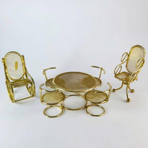May include: A miniature gold-colored patio set, including a round table and three chairs. The table and chairs have a mesh design and are made of metal. The set is designed for a dollhouse or miniature setting.