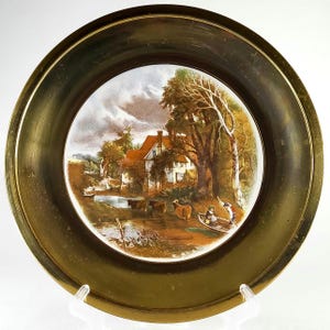 May include: A decorative plate with a landscape scene featuring a house with a red roof, trees, and a river. The plate has a gold-colored rim and a clear stand. The artwork depicts a rural setting with cows and people in a boat.