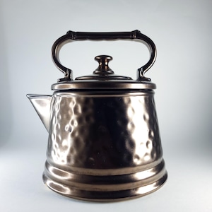 Puede incluir: Una tetera de color bronce con una textura martillada. La tetera tiene un asa curvada y un pequeño pico. La tapa está rematada con un pequeño pomo. La tetera está diseñada para su uso en la estufa.