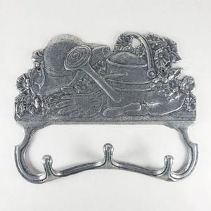 Può includere: Un appendiabiti da parete in metallo color argento con un design in rilievo raffigurante attrezzi da giardinaggio, tra cui un cappello da sole, un annaffiatoio, una cazzuola e guanti, circondati da motivi floreali. Ha tre ganci curvi.