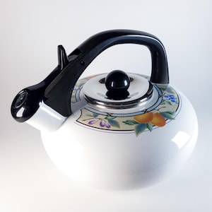 Op de afbeelding: Witte emaille theeketel met een zwarte handgreep en tuit. De ketel heeft een decoratieve band met fruit- en bloemmotieven. De deksel is verchroomd met een zwarte knop. Een klassiek keukenartikel.