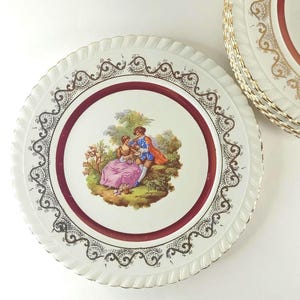 Puede incluir: Plato decorativo blanco con un borde burdeos y una ilustración detallada de una pareja en un jardín. El plato tiene un borde festoneado con ribete dorado y adornos ornamentados en negro y dorado. Platos adicionales apilados en el fondo.