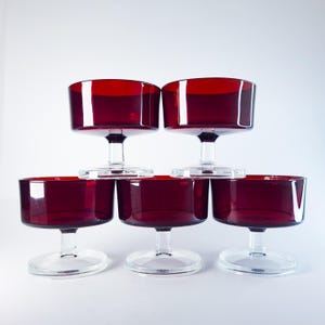 Può includere: Sei calici da champagne vintage in vetro rosso, impilati a piramide. I calici hanno una coppa ampia e poco profonda e un gambo corto con base circolare. Il colore rosso è intenso, su sfondo bianco.