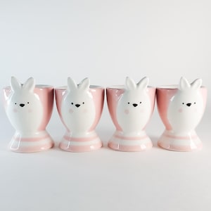 May include: Four ceramic egg cups featuring bunny designs. The egg cups are pink with white and pink striped bases. Each bunny has a white face, black eyes, and pink cheeks. The egg cups are arranged in a row.