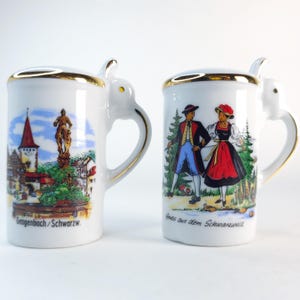 May include: Two white ceramic mugs with gold trim and handles. One mug features a painted scene of a town with a church and statue, the text "Gengenbach/Schwarzw." The other mug depicts people in traditional clothing, with the text "Gruss aus dem Schwarzwald."
