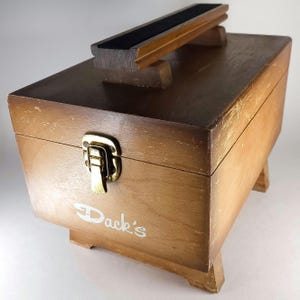 May include: A vintage wooden shoe shine box with a handle and a black felt pad. The box is rectangular with a hinged lid and a metal clasp. The text "Dack's" is visible on the front of the box.