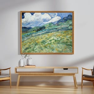 May include: A framed landscape painting with a wooden frame hangs above a light wood console table. The painting depicts a field with green and yellow grass, rolling hills, and a blue sky with white clouds. Two gray chairs flank the console.