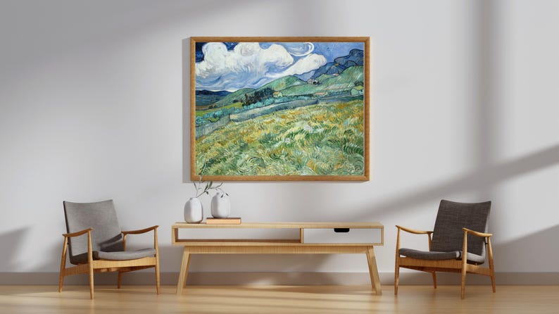 May include: A framed landscape painting with a wooden frame hangs above a light wood console table. The painting depicts a field with green and yellow grass, rolling hills, and a blue sky with white clouds. Two gray chairs flank the console.