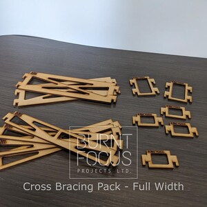 May include: A selection of laser-cut wooden cross bracing components. The image shows several long, angled pieces alongside smaller rectangular frames. The text reads "Cross Bracing Pack - Full Width".