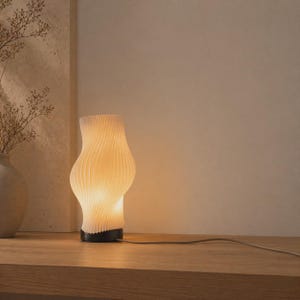 May include: A white, ribbed paper table lamp with a curved, hourglass shape. The lamp emits a warm glow, and sits on a dark base. A power cord extends from the base. The lamp is on a wooden surface.