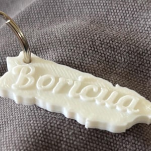 May include: A white keychain in the shape of Puerto Rico, with the word "Boriqua" in cursive script. The keychain is attached to a silver metal keyring.