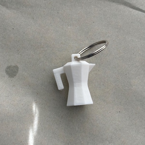 May include: A white, miniature coffee maker keychain with a silver key ring. The keychain is a small, detailed replica of a stovetop espresso maker, perfect for coffee lovers. The keychain is made of a hard, white material.