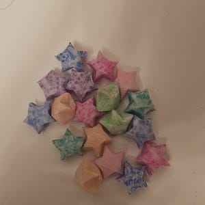 Puede incluir: Un conjunto de estrellas de la suerte de origami en varios colores, incluyendo rosa, azul, verde y naranja. Las estrellas est&aacute;n hechas de papel estampado y est&aacute;n dispersas sobre una superficie blanca. Algunas estrellas tienen texto.