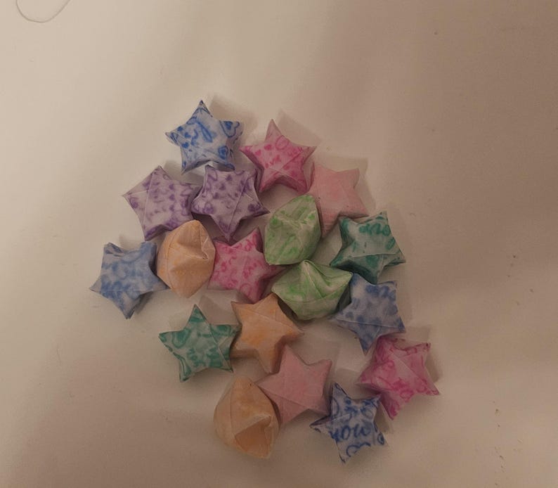 Puede incluir: Un conjunto de estrellas de la suerte de origami en varios colores, incluyendo rosa, azul, verde y naranja. Las estrellas est&aacute;n hechas de papel estampado y est&aacute;n dispersas sobre una superficie blanca. Algunas estrellas tienen texto.