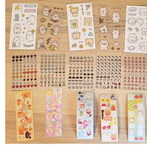 Könnte beinhalten: Eine Sammlung dekorativer Aufkleber mit verschiedenen Designs. Die Aufkleber zeigen niedliche Cartoon-Bären, Herzen und andere kleine Illustrationen. Die Farbpalette umfasst Weiß, Rosa, Gelb und andere Pastelltöne. Die Aufkleber sind auf einer Holzoberfläche angeordnet.