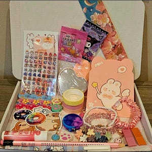 May include: A white box filled with stationery and accessories. Items include stickers, washi tape, a heart-shaped mirror, a pink bear-shaped card, bracelets, and pens. The box is open, revealing the contents.