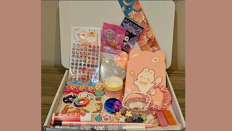 May include: A white box filled with stationery and accessories. Items include stickers, washi tape, a heart-shaped mirror, a pink bear-shaped card, bracelets, and pens. The box is open, revealing the contents.