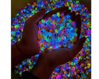 May include: A handful of colorful, glowing confetti in the shape of small flowers. The confetti is held in cupped hands, with a background of more confetti. The colors include blue, green, orange, and purple.