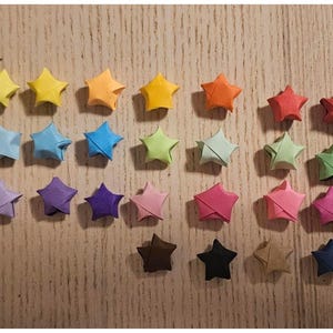 May include: A collection of colorful origami stars in various shades of yellow, orange, red, blue, green, pink, purple, brown, and black. The paper stars are arranged on a light brown wooden surface.