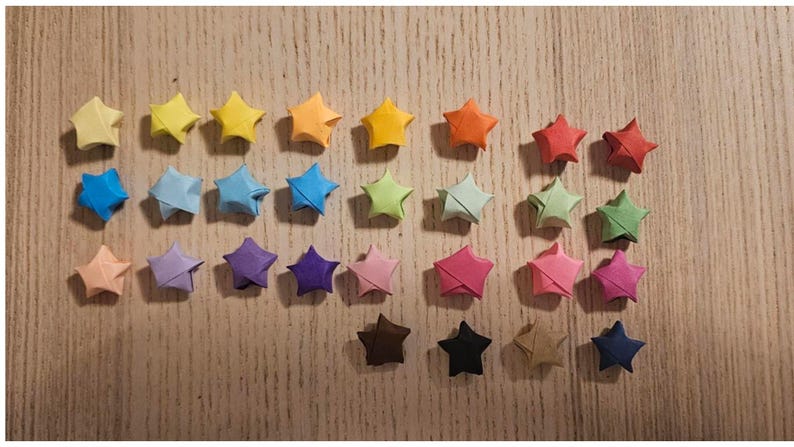 May include: A collection of colorful origami stars in various shades of yellow, orange, red, blue, green, pink, purple, brown, and black. The paper stars are arranged on a light brown wooden surface.