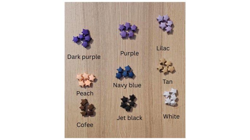 May include: A wooden surface displays small star-shaped origami in various colors: dark purple, purple, lilac, peach, navy blue, tan, jet black, coffee, and white. Text labels identify each color. The stars are grouped in small clusters.