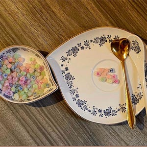 May include: A decorative serving set featuring a heart-shaped gold spoon and a white spoon. The set includes a floral patterned plate and a small dish, both filled with pastel-colored heart-shaped candies with letters.