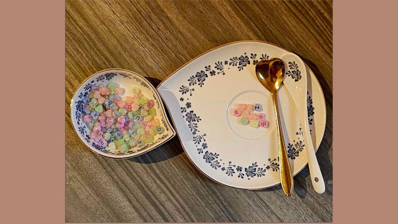 May include: A decorative serving set featuring a heart-shaped gold spoon and a white spoon. The set includes a floral patterned plate and a small dish, both filled with pastel-colored heart-shaped candies with letters.