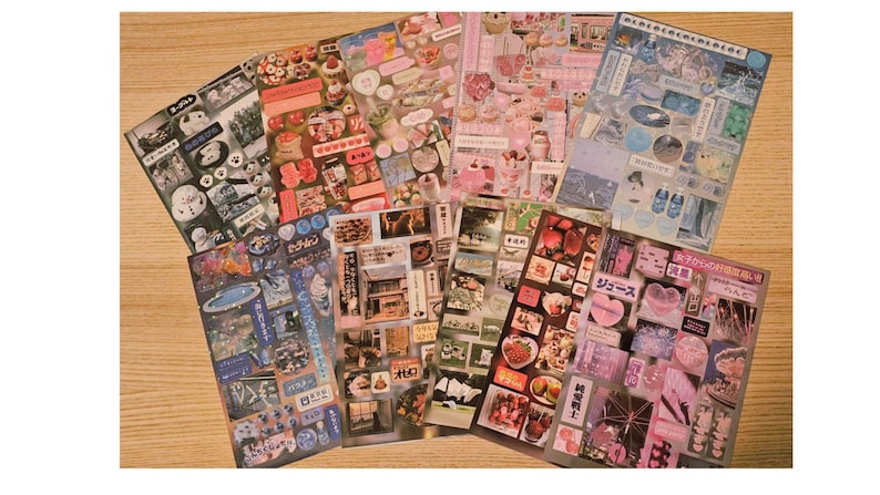 May include: A collection of decorative sticker sheets with various designs. The sheets feature images of food, desserts, and Japanese text. The color palette includes pink, white, and blue, with a wooden surface in the background.