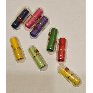 May include: A selection of colourful message capsules in clear plastic containers. The capsules are in various colours, including blue, pink, purple, green, red, and yellow. Each capsule has a gold-coloured band and a decorative element.