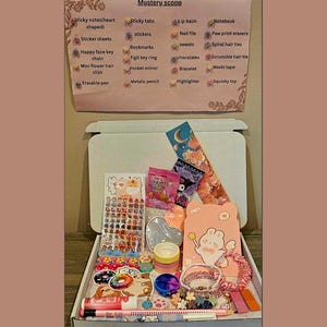 May include: An open white box filled with various stationery items, including stickers, pens, and a notebook. A sign above the box lists items such as sticky notes, stickers, and bookmarks. The items are in a variety of colors, including pink, blue, and yellow.