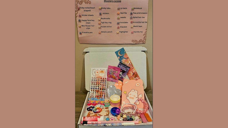 May include: An open white box filled with various stationery items, including stickers, pens, and a notebook. A sign above the box lists items such as sticky notes, stickers, and bookmarks. The items are in a variety of colors, including pink, blue, and yellow.