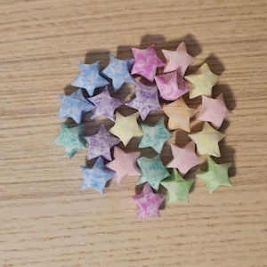 Puede incluir: Un conjunto de estrellas de origami en varios colores pastel, incluyendo azul, rosa, verde y amarillo. Las estrellas est&aacute;n dispuestas sobre una superficie de madera marr&oacute;n claro, creando una disposici&oacute;n visualmente atractiva. Algunas estrellas tienen texto.