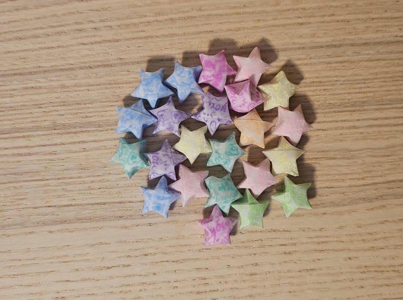Puede incluir: Un conjunto de estrellas de origami en varios colores pastel, incluyendo azul, rosa, verde y amarillo. Las estrellas est&aacute;n dispuestas sobre una superficie de madera marr&oacute;n claro, creando una disposici&oacute;n visualmente atractiva. Algunas estrellas tienen texto.