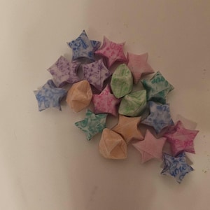 Puede incluir: Un conjunto de estrellas de origami en varios colores, incluyendo rosa, azul, morado, verde y melocot&oacute;n. Las estrellas est&aacute;n hechas de papel estampado y est&aacute;n dispuestas sobre una superficie blanca. Algunas estrellas tienen texto.