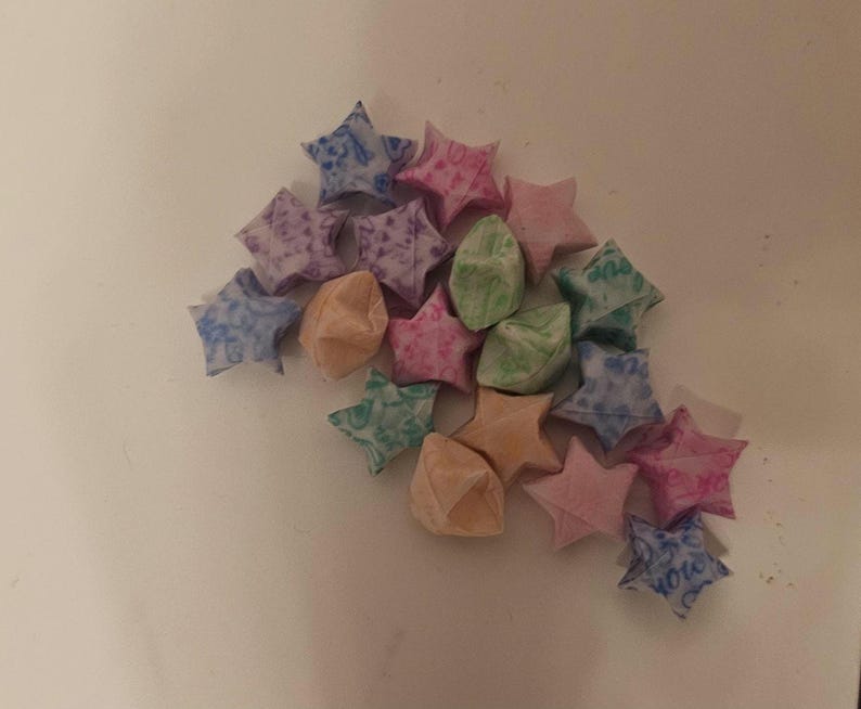 Puede incluir: Un conjunto de estrellas de origami en varios colores, incluyendo rosa, azul, morado, verde y melocot&oacute;n. Las estrellas est&aacute;n hechas de papel estampado y est&aacute;n dispuestas sobre una superficie blanca. Algunas estrellas tienen texto.