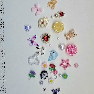 May include: A collection of small decorative charms and cabochons featuring various shapes including a teddy bear, a yellow rose, a red heart, a blue dolphin, a penguin, butterflies, bows, stars, flowers, and pearls in assorted colours and finishes.