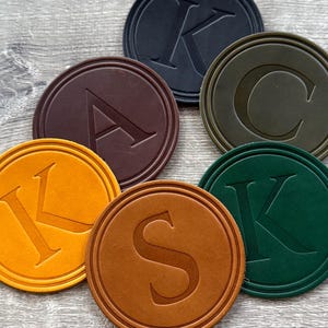 May include: A collection of six circular coasters in various colors including black, dark brown, olive green, mustard yellow, tan, and forest green. Each coaster features a debossed capital letter in the center, specifically K, A, C, K, S, and K.